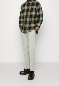 Chemise à carreaux aux teintes noires, vertes et jaunes; manches longues avec poignets à boutons. Associée à un pantalon vert clair et des chaussures noires à pampilles.