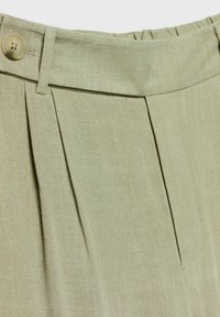 Gros plan de la ceinture d’un pantalon beige clair avec bouton, passants de ceinture et plis avant montrant la texture du tissu.