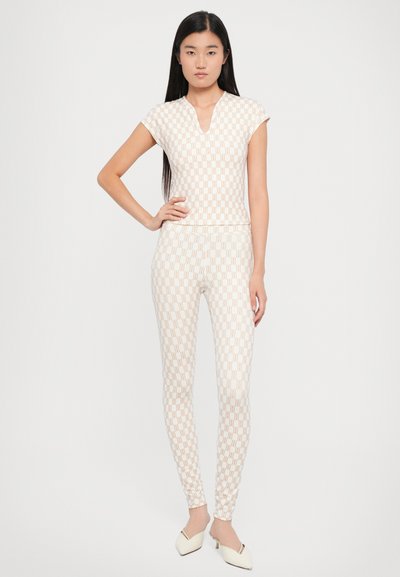 Witte stoffen jumpsuit met een geometrisch patroon in beige. Heeft een v-hals, korte mouwen en aansluitende pijpen, gecombineerd met witte hakken.