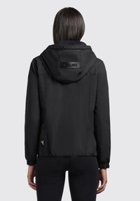 Veste légère noire avec capuche, dotée de poignets élastiques, d'une texture lisse et d'un petit patch logo sur le côté inférieur.