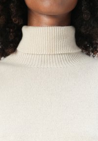 Beige Rollkragenpullover mit gerippter Textur und tailliertem Design, ausgestattet mit einem hohen umgeschlagenen Kragen und einem glatten Strickkörper.