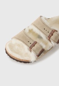 Beige semsket skinnsandaler med to justerbare stropper, foret med hvit kunstpels, og brun korksåle med subtile merkevarerdetaljer.