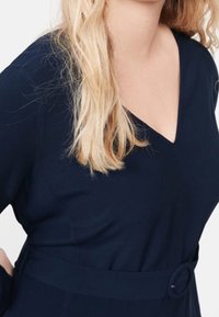 Navy mekko, jossa on V-pääkallo, pitkähihaiset hihat ja vyötäröllä vyö, jossa on pyöreä solki. Sujuva kangas ja istuva yläosa.