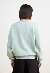 Persona con pelo negro corto que lleva un suéter de gran tamaño de color azul claro y pantalones negros, de espaldas contra un fondo blanco plano.