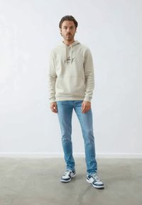 Fato de capuz bege claro com um bolso frontal e logótipo, combinado com jeans azul claro e sapatilhas pretas e brancas com detalhes azuis. Fundo simples.