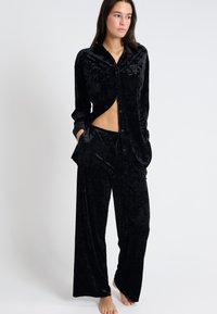 Conjunto de pijama de terciopelo negro que incluye una camisa de botones con cuello y mangas largas, acompañada de pantalones de pierna ancha. Textura suave y ajuste relajado.