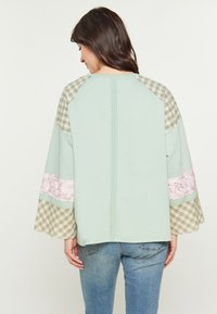 Menta zöld, könnyű felső, kockás ujjal és virágos díszítéssel. Lazább szabású és bordázott nyakkivágása van. Hátsó varrás részletek láthatók.