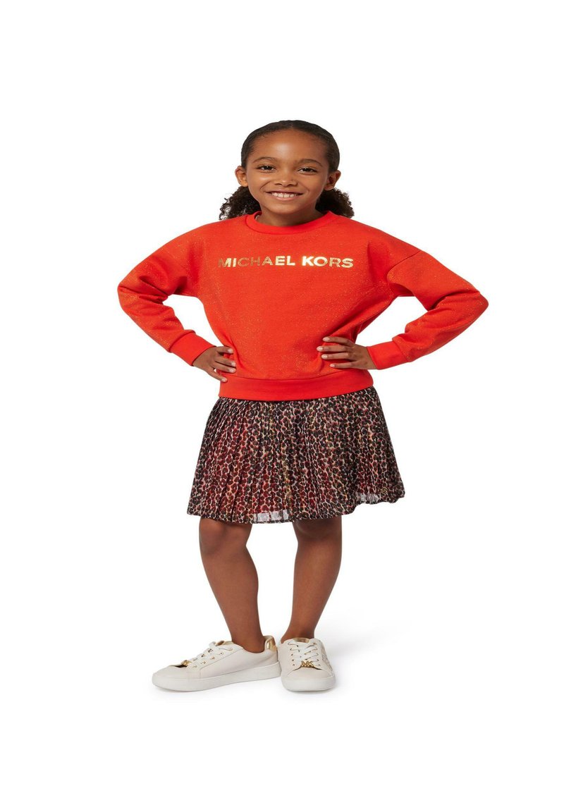 Sweatshirt rouge avec "MICHAEL KORS" en doré, associé à une jupe plissée avec un imprimé animal noir et marron. Baskets blanches avec des accents dorés.
