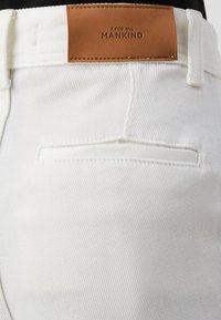 Witte denim jeans met een achterzak en een tan leren taillebandlabel waarop "7 FOR ALL MANKIND" is gedrukt. Textuurstof met zichtbare stiksels.