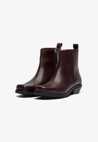 Botas de cuero burdeos hasta el tobillo con punta redondeada, cremallera lateral y tirador, que presentan un tacón bajo apilado y una textura suave.