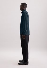 Camicetta a maniche lunghe di un verde scuro con polsini a bottoni, abbinata a pantaloni e scarpe nere. Linee pulite e una vestibilità su misura con una texture morbida.