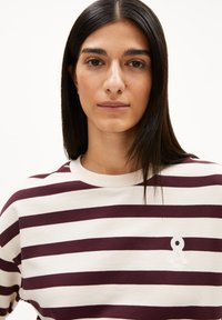 Junge Frau mit geradem, dunklem Haar, die ein weißes und kastanienbraunes gestreiftes Shirt mit einem kleinen weißen Logo auf der Brust trägt, vor einem einfarbigen Hintergrund.