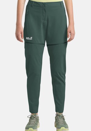 Pantaloni da trekking verde scuro con zip staccabile, tasche frontali e logo sulla coscia sinistra, indossati con scarpe da trekking verdi su una persona in piedi.