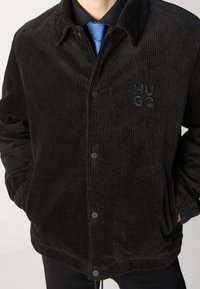 Homme portant une veste en velours côtelé noire avec « HUGO » brodé sur la poitrine, cravate bleue et chemise noire, les mains dans les poches de la veste.