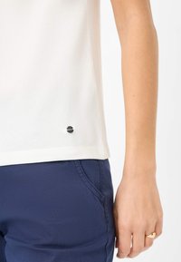 Witte top met korte mouwen en een ronde zoom, voorzien van een klein zilver logo accent nabij de onderrand, gecombineerd met marineblauwe broek.