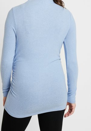 Femme portant un haut ajusté à manches longues bleu clair et un pantalon noir, vue de dos sur un fond blanc uni.