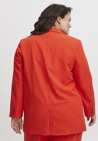 Oranžinė švarkas su pritaikytu siluetu, įpjovomis apykaklėmis ir užsegamais rankovėmis, su centriniu ventiliaciniu plyšiu nugaroje, kad būtų lengviau judėti.