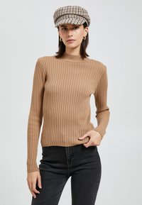 Brązowy sweter z długim rękawem i wysokim kołnierzem. W połączeniu z czarnymi dżinsami, kaszkietem w wzór houndstooth oraz kolczykami w kształcie kół.