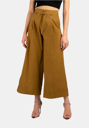 Pantaloni larghi marroni con pieghe, vita alta, cintura annodata, texture liscia e fondo a risvolto, abbinati a sandali con tacco alto e cinturini.