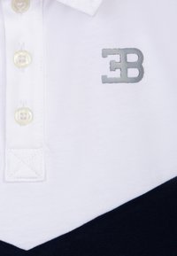 Polo shirt bianco con design a zigzag blu navy, colletto con tre bottoni e logo ricamato in argento sul lato sinistro del petto. Materiale in cotone morbido.