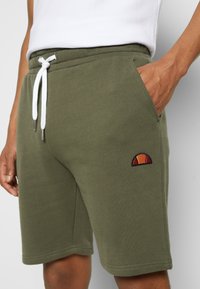 Pantaloni corti verde oliva realizzati in tessuto morbido, dotati di tasche laterali, una vita elasticizzata con lacci bianchi e un piccolo logo ricamato.