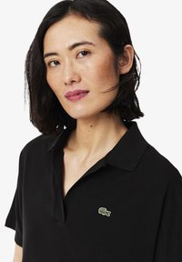 Svart polo-shirt i slät tyg, med klassisk krage och korta ärmar. Anmärkningsvärt grönt krokodil-logotyp på bröstet.