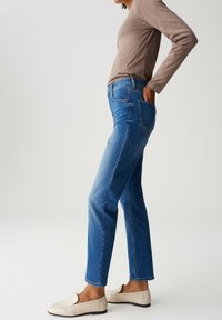 Vrouw in een blauwe jeans, beige lange mouwen top en crèmekleurige instappers, staande in profiel met één hand in de achterzak.