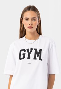 Biele tričko s krátkym rukávom so suverénnym čiernym nápisom "GYM" cez hruď a malým logom pod ním. Mäkká bavlnená látka, uvoľnený strih.