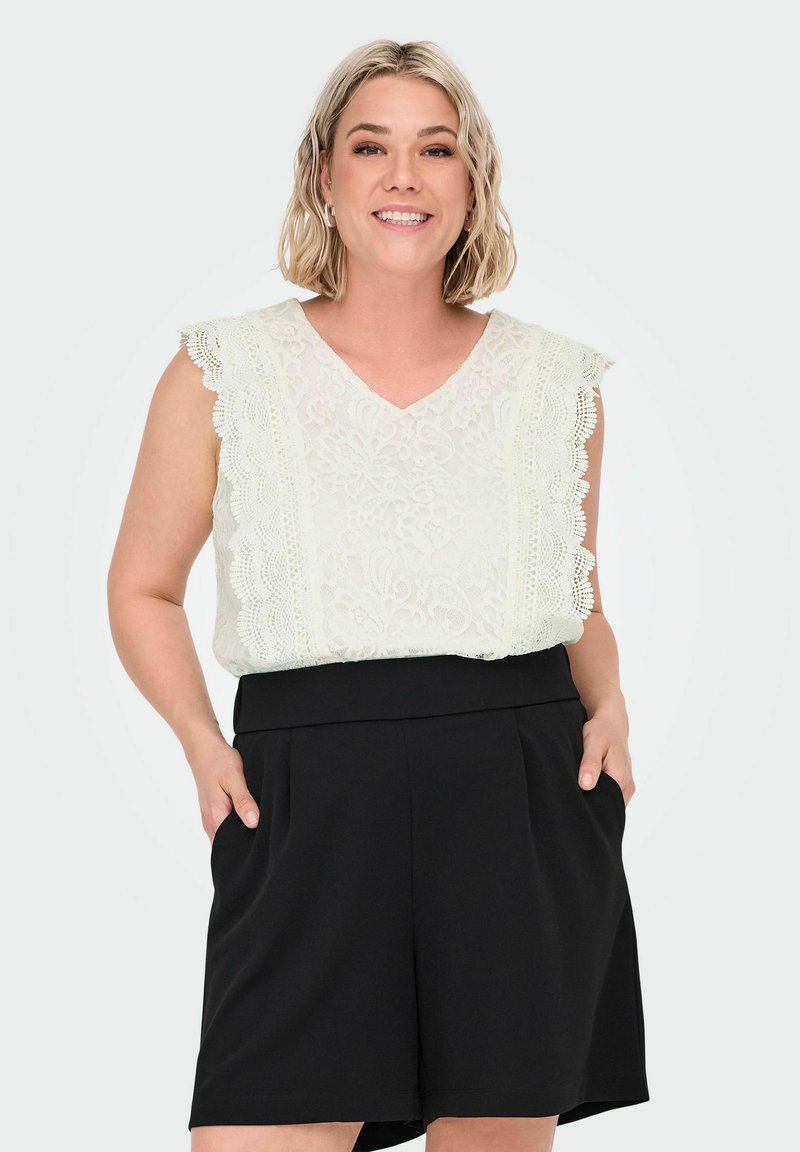 Smilende kvinde med kort blont hår iført en ærmeløs hvid blonde top og højtaljede sorte shorts med hænderne i lommerne.