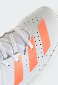 Hvid atletiksko med tre orange striber, tekstureret overdel, perforeringer for åndbarhed og hvide snørebånd. Gummisålens detalje er synlig.
