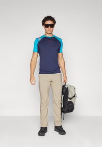 Ανδρικό αθλητικό μπλούζα μπλε και ναυτική με κοντά μανίκια, μπεζ cargo παντελόνι, μαύρα αθλητικά παπούτσια, κρατώντας ένα γκρι και μαύρο σακίδιο, με γυαλιά ηλίου στο πρόσωπο.