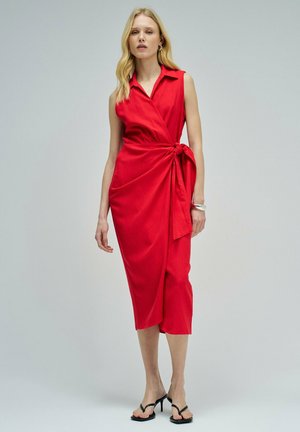 Mujer con cabello largo y rubio, vistiendo un vestido rojo sin mangas con lazada lateral y sandalias negras de tacón alto sobre fondo gris.