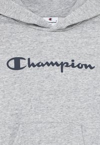 Grå hettegenser laget av mykt stoff, med en frontlomme og en marineblå "Champion"-logo på brystet. Stoffet har et litt teksturert utseende.
