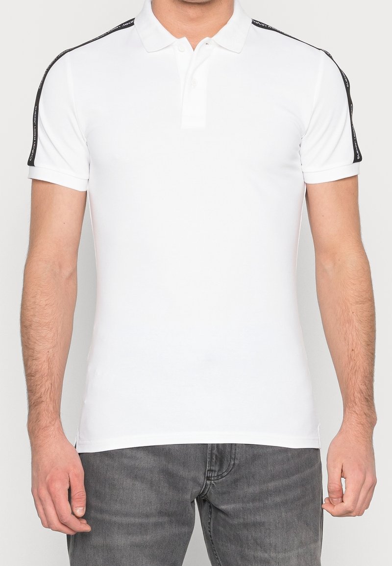 Homme portant un polo blanc à manches courtes avec des bandes noires de marque sur les épaules et un jean gris, debout devant un fond uni.