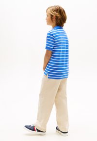 Αγόρι που στέκεται πλαγίως, φορώντας μπλε και λευκή ριγέ πόλο, μπεζ παντελόνι και μπλε ναυτικό αθλητικά παπούτσια με κόκκινες και λευκές ρίγες.