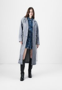 Long manteau en denim gris avec fermeture à boutons, poches latérales et fentes. Porté par-dessus une robe chemise en denim bleu et des bottes noires jusqu'aux genoux.