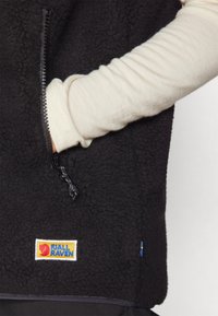 Schwarze Fleece-Weste aus strukturiertem Material, mit Reißverschlusstasche und Fjällräven-Logo. Darunter trägt die Person ein langärmliges, helles Oberteil.
