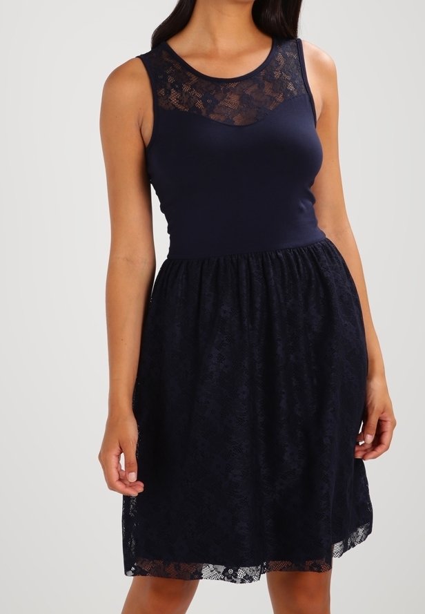 Femme portant une robe sans manches bleu marine avec un détail en dentelle sur le haut de la poitrine et une jupe superposée en dentelle, debout devant un fond uni.