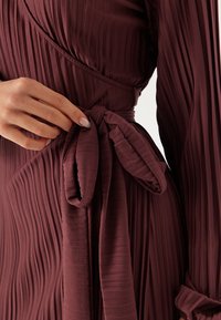 Robe plissée mauve à manches longues, dotée d'une ceinture nouée à la taille. Le tissu a un design texturé et côtelé.