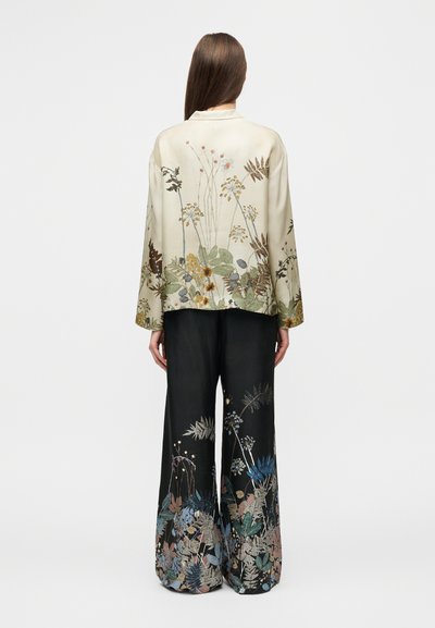 Veste en soie beige avec broderies florales, associée à un pantalon large noir orné de motifs botaniques colorés à l'ourlet.
