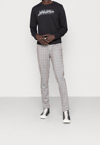 Maglietta nera a maniche lunghe con stampa grafica bianca, abbinata a pantaloni grigi a quadri slim-fit e sneakers nere con suola bianca.