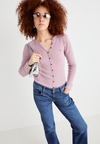 Haut mauve clair à manches longues en mailles avec détails de boutons, associé à un jean en denim. Le modèle tient un sac argenté métallique et porte des lunettes de soleil.