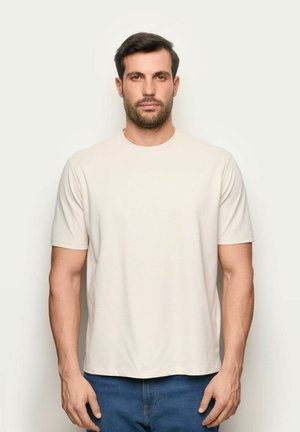 Uomo con capelli scuri corti e barba che indossa una t-shirt beige a maniche corte e jeans blu, in piedi davanti a uno sfondo bianco.