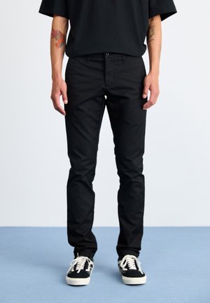 Pantalons en coton noir à coupe slim, avec une fermeture à bouton à l'avant et cinq poches, associés à des baskets noir et blanc.