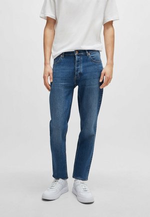 Homme portant un jean bleu droit, un t-shirt blanc et des baskets blanches, debout devant un fond clair uni.