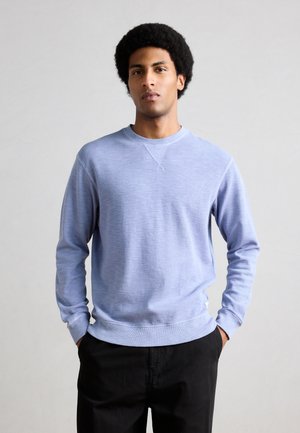 Junger Mann mit lockigem Haar, der ein hellblaues Langarmhemd und schwarze Hosen trägt und mit den Händen in den Taschen vor einem grauen Hintergrund steht.