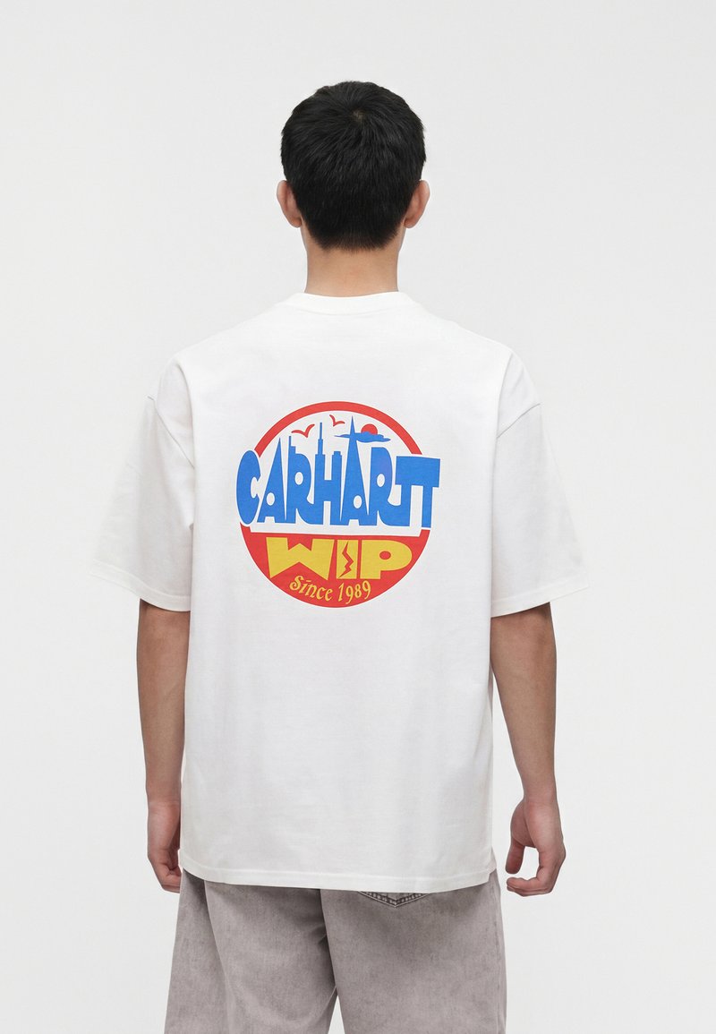 Uomo che indossa una maglietta bianca con un grande logo circolare Carhartt WIP in blu, rosso e giallo sulla schiena, in piedi davanti a uno sfondo bianco semplice.