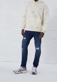 Sudadera crema con cordones, bolsillo y detalles de costura; vaqueros azules desgastados; zapatillas deportivas grises y doradas. Fondo sencillo.