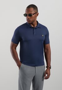 Granatowa koszulka polo z krótkimi rękawami, gładką fakturą, kołnierzykiem na trzy guziki oraz trójkątną naszywką z logo na lewej piersi, zestawiona z szarymi spodniami.