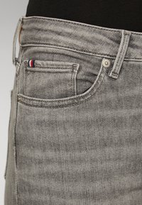 Acercamiento de un bolsillo de jeans de mezclilla gris con remaches de metal y una pequeña etiqueta de tela roja, blanca y azul en el bolsillo para monedas.